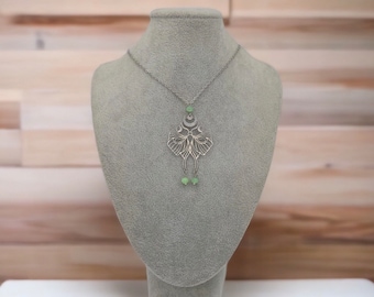 Op de afbeelding: Een zilveren ketting met een delicate, ingewikkelde hanger in de vorm van een mantarog. De hanger is versierd met kleine, lichtgroene edelstenen. De ketting wordt getoond op een grijze fluwelen buste.