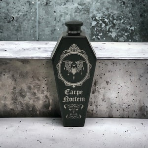 May include: A matte black flask shaped like a coffin with a screw-top lid. The front features an engraved circular frame containing a bat illustration with a crescent moon and stars, with the text Carpe Noctem printed in a gothic font below.