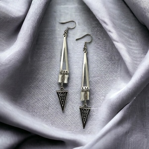 Puede incluir: Un par de pendientes de plata con un colgante triangular y una cadena de cuentas colgantes. Los pendientes est&aacute;n sobre un fondo de tela gris.
