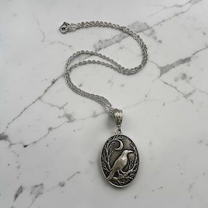 Puede incluir: Un collar de metal plateado con un colgante ovalado que presenta un dise&ntilde;o en relieve de un cuervo posado sobre una rama bajo una luna creciente. El colgante cuelga de una delicada cadena plateada con un cierre de mosquet&oacute;n.