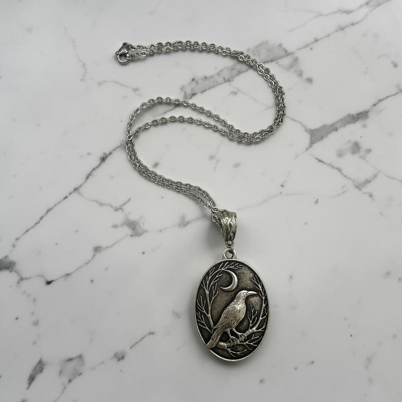 Puede incluir: Un collar de metal plateado con un colgante ovalado que presenta un dise&ntilde;o en relieve de un cuervo posado sobre una rama bajo una luna creciente. El colgante cuelga de una delicada cadena plateada con un cierre de mosquet&oacute;n.
