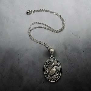 Puede incluir: Un collar con colgante ovalado de tono plateado que presenta un relieve detallado de un cuervo posado en una rama bajo una luna creciente. El colgante cuelga de una delicada cadena plateada con una anilla texturizada.