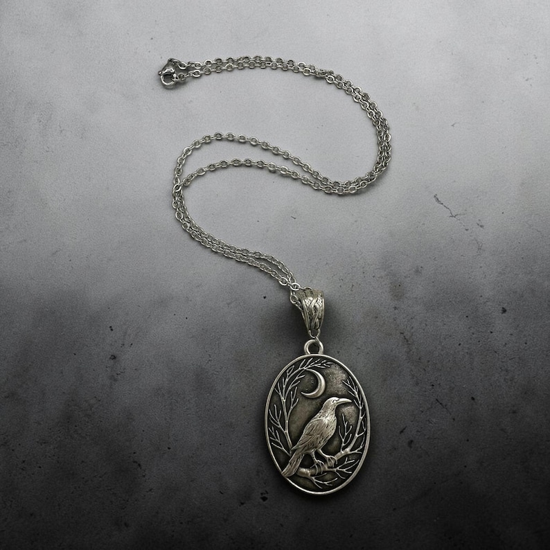 Puede incluir: Un collar con colgante ovalado de tono plateado que presenta un relieve detallado de un cuervo posado en una rama bajo una luna creciente. El colgante cuelga de una delicada cadena plateada con una anilla texturizada.