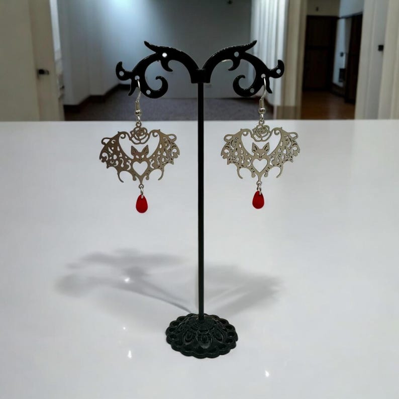 May include: Silver bat earrings with red teardrop crystals hanging from the bottom. The earrings are displayed on a black stand.