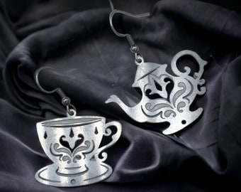 May include: A pair of silver earrings featuring a teapot and a teacup design. The earrings are intricate and detailed, with a delicate floral pattern.