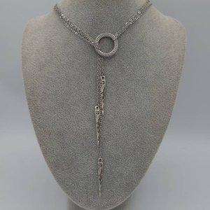 May include: A silver-toned lariat necklace featuring a central circular ring with a Greek key pattern. Three delicate chains hang from the ring, each ending in a slender, pointed metal pendant with intricate cut-out detailing and a metallic finish.