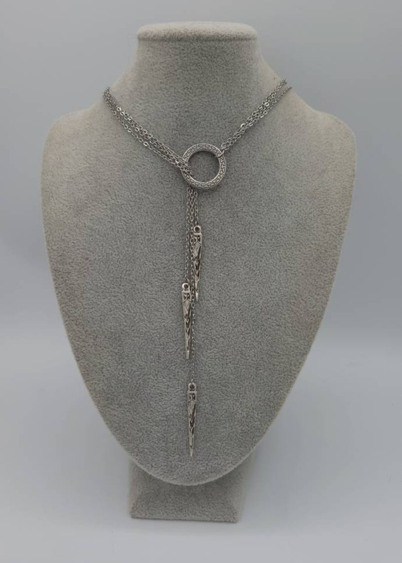 May include: A silver-toned lariat necklace featuring a central circular ring with a Greek key pattern. Three delicate chains hang from the ring, each ending in a slender, pointed metal pendant with intricate cut-out detailing and a metallic finish.