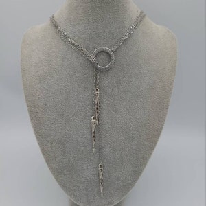 May include: A silver-toned lariat necklace featuring a central circular ring with a Greek key pattern. The necklace has multiple delicate chains that drape downwards, ending in two elongated, pointed metal pendants with intricate engraved detailing.