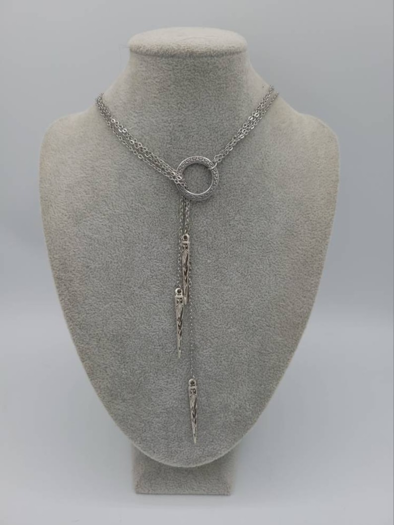 May include: A silver-toned lariat necklace featuring a central circular ring with a Greek key pattern. The necklace has multiple delicate chains that drape downwards, ending in two elongated, pointed metal pendants with intricate engraved detailing.