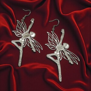 May include: A pair of silver-colored earrings featuring a skeleton fairy design. The earrings have detailed skeletal structures, delicate wing shapes, and are suspended from simple ear wires. The background is a deep red, textured fabric.