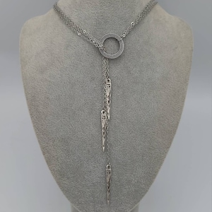May include: A silver-toned lariat necklace featuring a central circular ring with a textured Greek key pattern. Three delicate chains hang from the ring, each ending in a slender, pointed metal pendant with intricate engraved detailing.