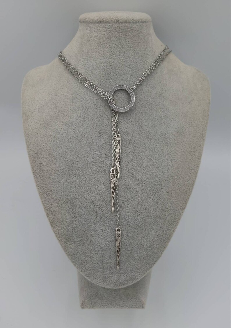 May include: A silver-toned lariat necklace featuring a central circular ring with a textured Greek key pattern. Three delicate chains hang from the ring, each ending in a slender, pointed metal pendant with intricate engraved detailing.