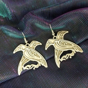 May include: A pair of silver earrings with a detailed design of a bird. The earrings are on a dark blue and purple iridescent fabric.