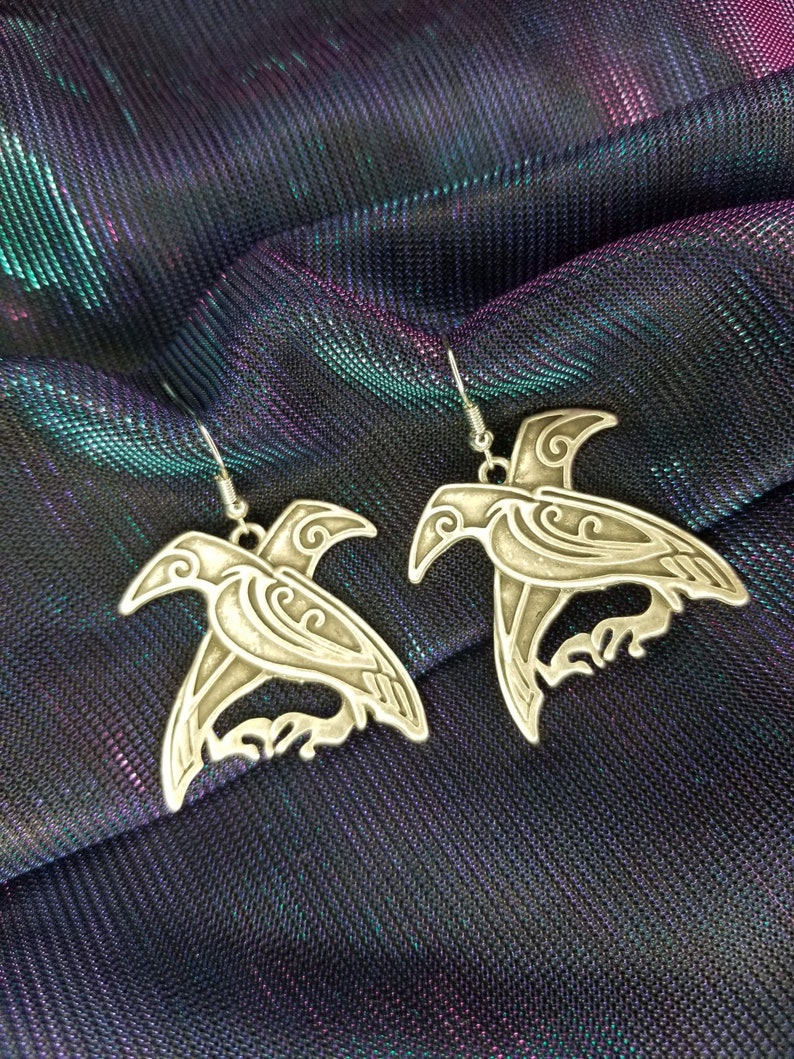 May include: A pair of silver earrings with a detailed design of a bird. The earrings are on a dark blue and purple iridescent fabric.