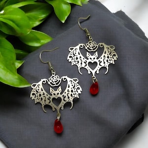May include: A pair of silver earrings shaped like bats with a red glass drop hanging from each. The bats have a heart-shaped body with a rose design on the top.