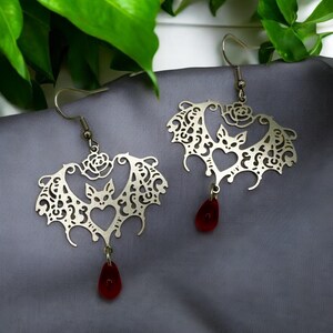 May include: A pair of silver earrings shaped like bats with intricate cutouts and red glass teardrop dangles.