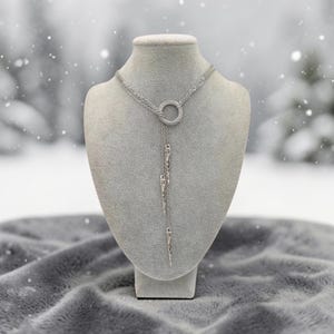 May include: A silver-toned lariat necklace featuring a central circular ring connector. Three delicate chains extend downward from the ring, each terminating in a slender, pointed metallic spike pendant with a textured, etched surface finish.