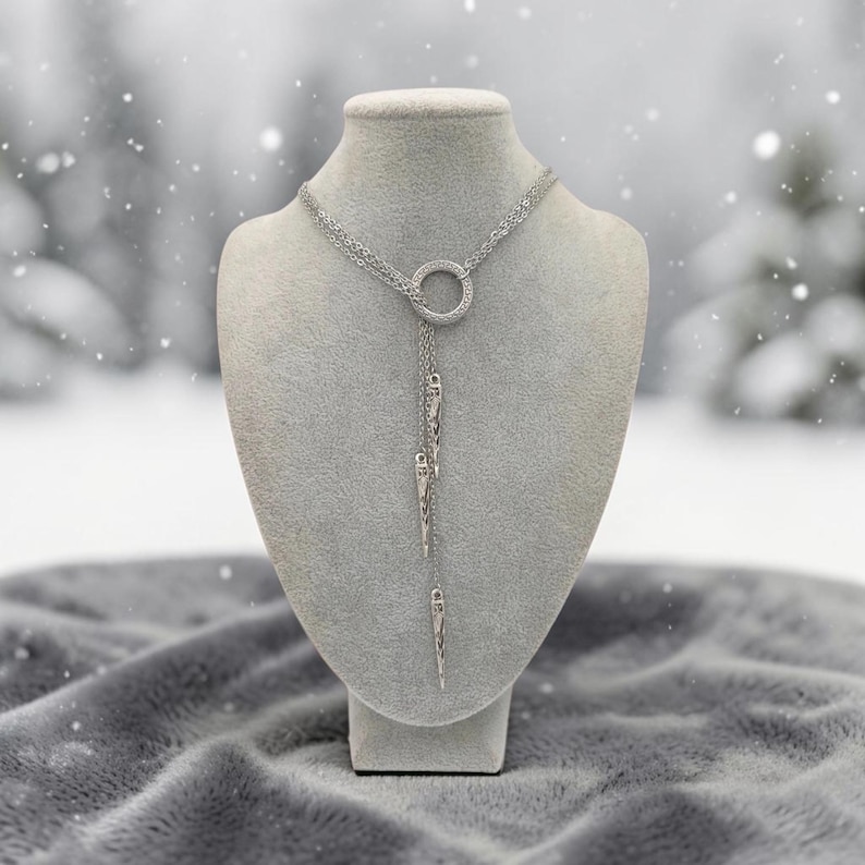 May include: A silver-toned lariat necklace featuring a central circular ring connector. Three delicate chains extend downward from the ring, each terminating in a slender, pointed metallic spike pendant with a textured, etched surface finish.