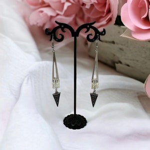 Puede incluir: Un par de pendientes colgantes de plata con detalles negros y un dise&ntilde;o triangular. Los pendientes est&aacute;n colgados de un soporte de pendientes negro.