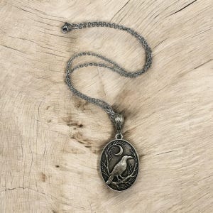 Puede incluir: Un collar con colgante ovalado de tono plateado que presenta un relieve detallado de un cuervo posado en una rama bajo una luna creciente. El colgante cuelga de una delicada cadena plateada a juego con un acabado oxidado y texturas.