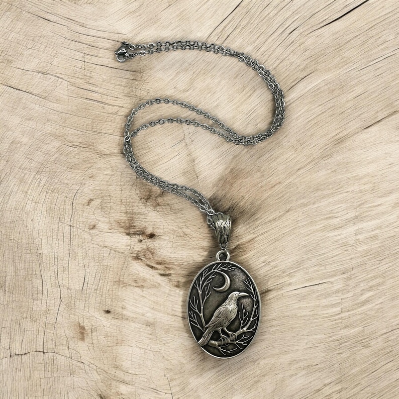 Puede incluir: Un collar con colgante ovalado de tono plateado que presenta un relieve detallado de un cuervo posado en una rama bajo una luna creciente. El colgante cuelga de una delicada cadena plateada a juego con un acabado oxidado y texturas.