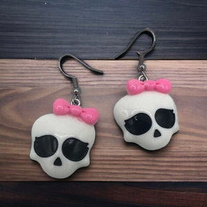 May include: A pair of white skull earrings with pink bows. The earrings are made of a sparkly material and have black eyes.