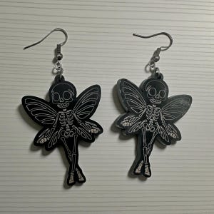 May include: A pair of black skeleton fairy earrings. Each earring features a skeletal fairy design with detailed white outlines on the wings and body. The earrings have silver-colored fish hook closures.