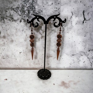 May include: A pair of dangle earrings with a bronze finish. The earrings feature three round beads with a swirl design and a pointed drop.
