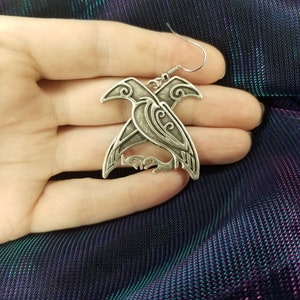 May include: Silver earrings featuring a detailed design of two intertwined birds with intricate patterns.