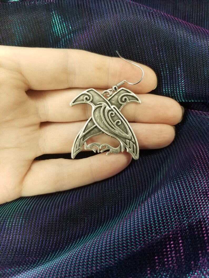 May include: Silver earrings featuring a detailed design of two intertwined birds with intricate patterns.