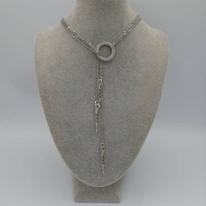 May include: A silver-toned lariat necklace featuring a central circular ring encrusted with small clear stones. Two delicate chains extend downward from the ring, each terminating in a slender, pointed metal pendant with etched detailing.