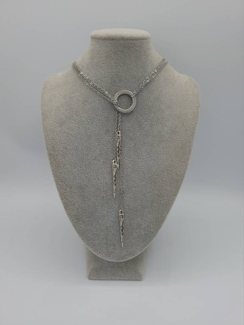 May include: A silver-toned lariat necklace featuring a central circular ring encrusted with small clear stones. Two delicate chains extend downward from the ring, each terminating in a slender, pointed metal pendant with etched detailing.