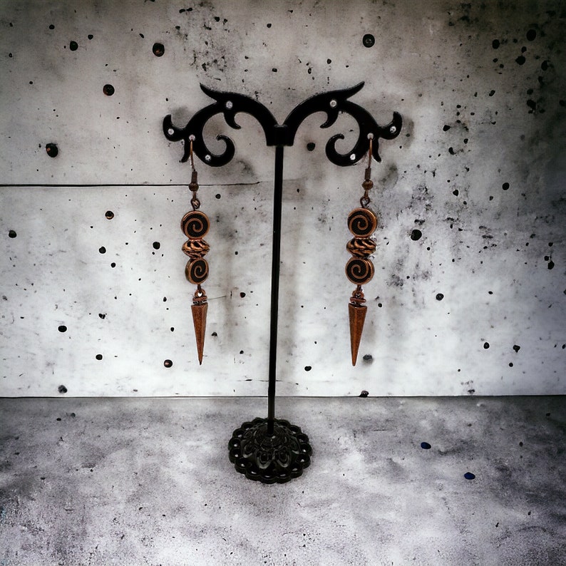 May include: A pair of dangle earrings with a copper finish. The earrings feature three round beads with a swirl design and a pointed drop.