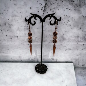 May include: A pair of long, copper-toned earrings with a spiral design hang from a black stand. The earrings have a pointed tip and are made of metal.