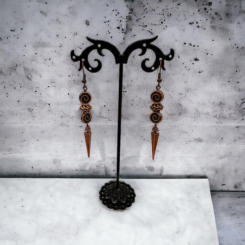 May include: A pair of long, copper-toned earrings with a spiral design hang from a black stand. The earrings have a pointed tip and are made of metal.