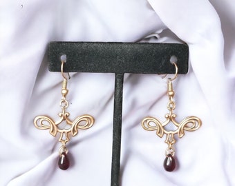May include: A pair of gold-toned metal earrings featuring a symmetrical, scrollwork design with a central vertical element. Each earring hangs from a hook wire and terminates in a small, dark red, teardrop-shaped gemstone bead suspended at the bottom.