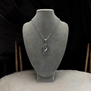 Puede incluir: Un collar con colgante ovalado de tono plateado que presenta un dise&ntilde;o en relieve de un cuervo posado en una rama bajo una luna creciente. El colgante cuelga de una delicada cadena plateada, mostrando detalles intrincados y un acabado met&aacute;lico.