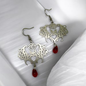 May include: Silver earrings with a bat design and a red glass drop. The bat is surrounded by a delicate filigree pattern.