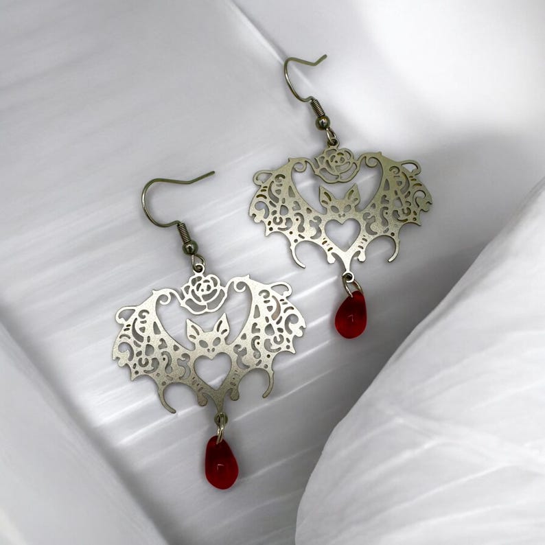 May include: Silver earrings with a bat design and a red glass drop. The bat is surrounded by a delicate filigree pattern.