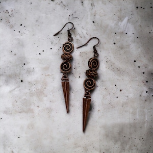 May include: A pair of long, copper earrings with a pointed, triangular pendant. Each earring features three copper beads with a black swirl design.
