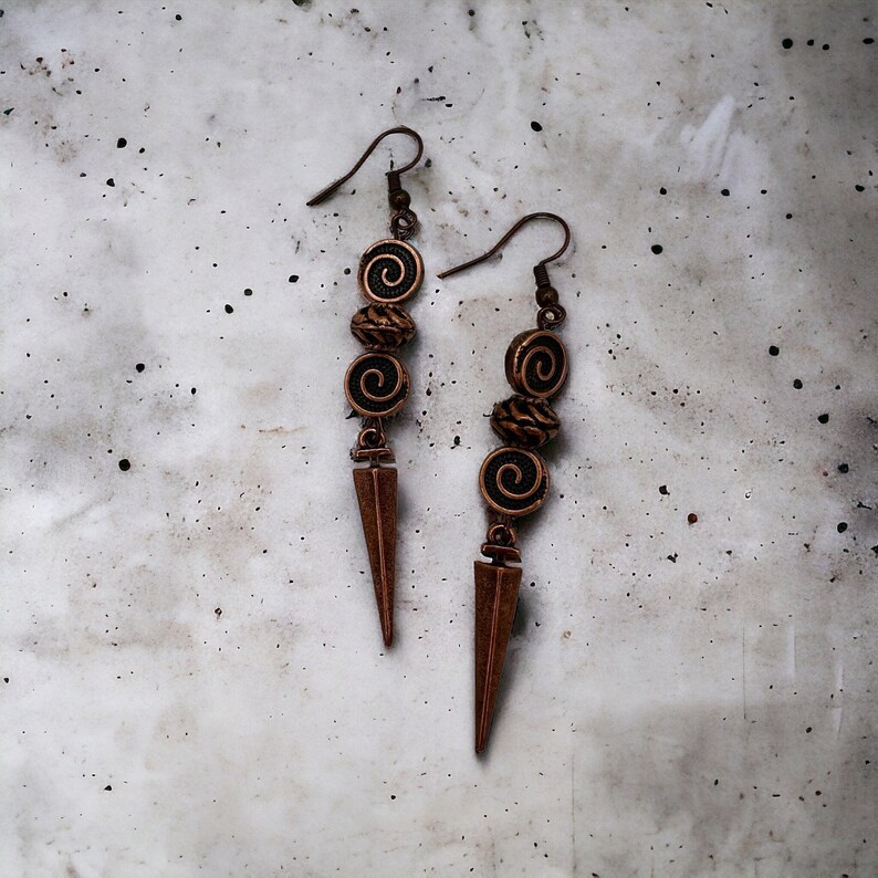 May include: A pair of long, copper earrings with a pointed, triangular pendant. Each earring features three copper beads with a black swirl design.