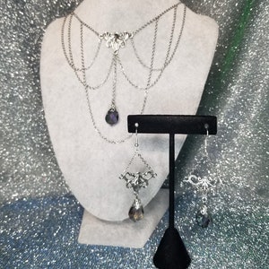 May include: A silver necklace with a dangling crystal pendant and matching earrings. The necklace has a delicate, intricate design and features multiple chains.