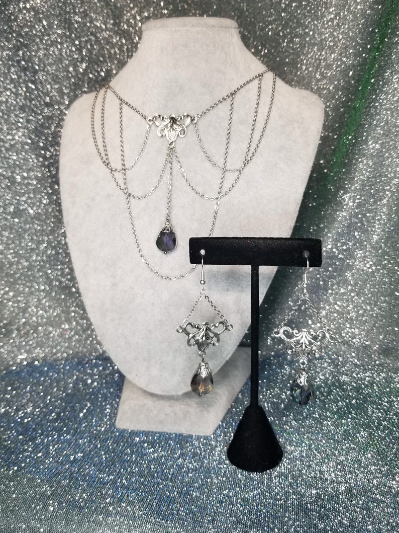 May include: A silver necklace with a dangling crystal pendant and matching earrings. The necklace has a delicate, intricate design and features multiple chains.