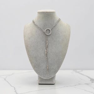 May include: A silver-toned lariat necklace featuring a central circular ring connector. Three delicate chains extend downward from the ring, each terminating in a slender, pointed metal spike pendant. The design is symmetrical and metallic.