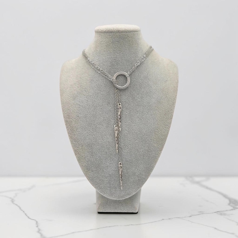 May include: A silver-toned lariat necklace featuring a central circular ring connector. Three delicate chains extend downward from the ring, each terminating in a slender, pointed metal spike pendant. The design is symmetrical and metallic.