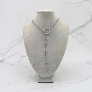 May include: A silver-toned lariat necklace featuring a central circular ring connector. Three delicate chains extend downward from the ring, each terminating in a slender, pointed metal spike pendant. The necklace has a polished metallic finish.