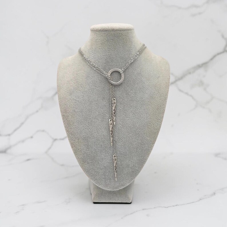 May include: A silver-toned lariat necklace featuring a central circular ring connector. Three delicate chains extend downward from the ring, each terminating in a slender, pointed metal spike pendant. The necklace has a polished metallic finish.