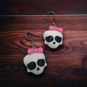 May include: A pair of white skull earrings with black eyes and pink bows. The earrings are made of metal and have a hook closure.