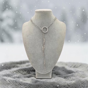 May include: A silver-toned lariat necklace featuring a central circular ring with a textured surface. Three delicate chains of varying lengths hang from the ring, each ending in a slender, pointed, metallic spike-shaped pendant.