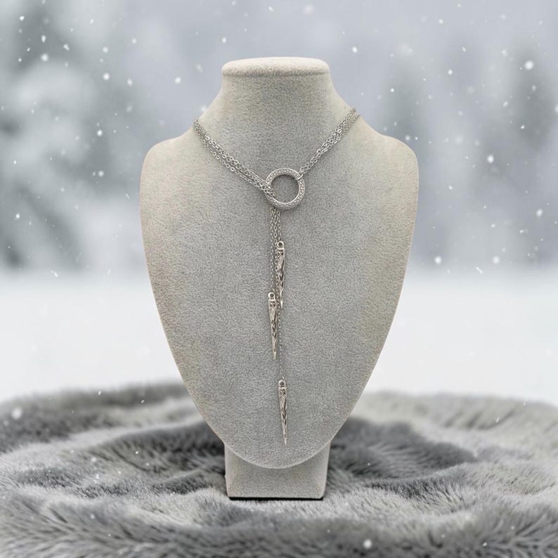 May include: A silver-toned lariat necklace featuring a central circular ring with a textured surface. Three delicate chains of varying lengths hang from the ring, each ending in a slender, pointed, metallic spike-shaped pendant.